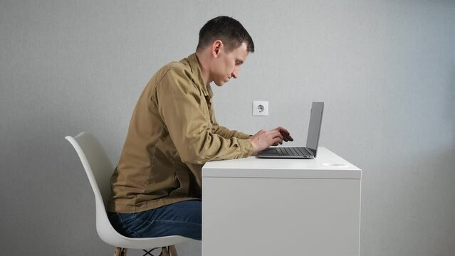 Young man fidgets on chair working on laptop against grey wall. Employee in mustard jacket feels uncomfortable sitting at table in office side view