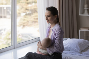 Smiling dreamy young mother looking in distance while breastfeeding little newborn baby boy girl, imagining future of infant son daughter, enjoying happy motherhood, resting on bed in modern bedroom.