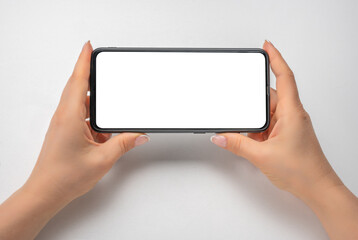 phone mockup. female hands hold phone horizontally. cell phone in horizontal position with blank white screen on gray table. Woman holding mobile cellphone in horizontal with isolated screen
