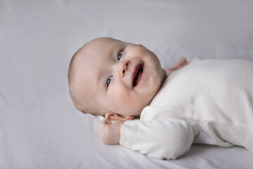 Head shot close up happy small cute kid boy girl in white bodysuit lying on bed, laughing looking...
