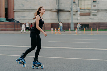 Fototapeta premium Athletic woman on roller skates moves actively poses on asphalt at street dressed in active wear has cheerful expression