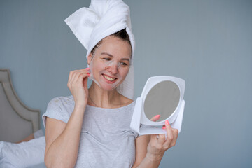 Pretty woman in white towel is relaxing in bedroom and doing moisturizing and restorative facial mask near mirror. Girl doing beauty treatment and relaxing at home. Morning skin care beauty routine