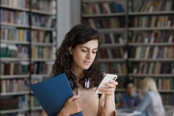 Student girl hold smartphone standing in library, take break between lessons, check timetable online, use mobile application, chat in social media. Generation Z, modern tech usage, lifestyle concept