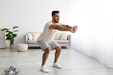 Muscular young Arab man doing squats, working out at home, copy space