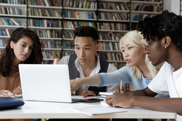 Four multiethnic schoolmates look at laptop discuss online task, learning use internet resources, search information busy in studying sit at table in university library. Higher education, tech concept