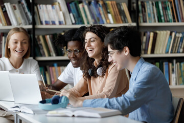 Laughing multi ethnic students use laptop studying or having fun using laptop sit at shared table in university library, having friendly relations, prepare for exams together using modern tech concept