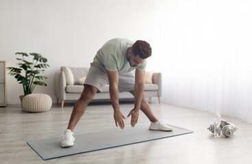 Fit young Arab man doing exercises, stretching legs muscles at home, full length