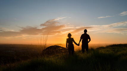 Fototapeta na wymiar Silhouette couple standing on the hilltop looking at the sun rising over Auckland city. Photo taken at One Tree Hill.