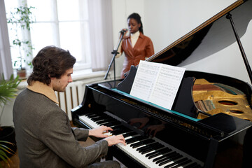 Young pianist sitting in front of the sheet music and playing the music on piano with African woman...