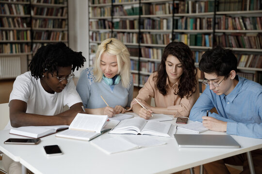 Four Multi Ethnic Students Take Notes Writing In Copybook While Studying, Prepare For Exams Seated At Table In University Library Looking Concentrated Busy In High School Assignment. Education Concept