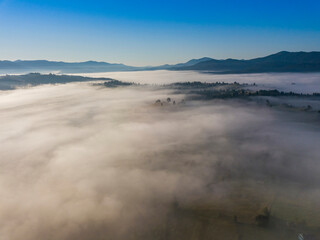 Fototapeta na wymiar Flight over fog in Ukrainian Carpathians in summer. Mountains on the horizon. A thick layer of fog covers the mountains with a continuous carpet. Aerial drone view.
