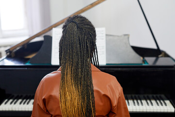 Rear view of young woman with stylish hairstyle playing the grand piano during lesson at musical...