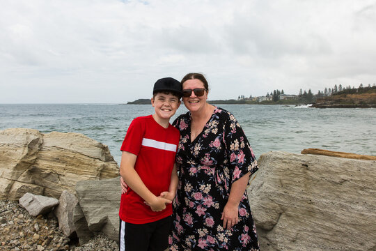 Mother and son standing together smiling on rock wall in front of beach water