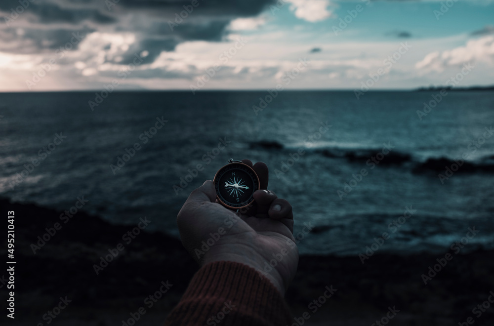 Wall mural Travel adventure lifestyle concept with man holding navigation compass in front of dramatic sky and ocean landscape. Journey and destination. Lost direction and life. People finding the way