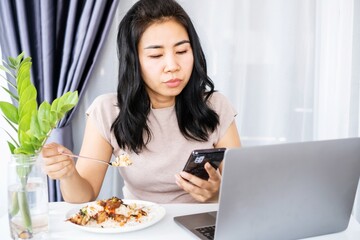 busy Asian businesswoman eating, having meal and working at office with laptop on desk hand holding mobile phone