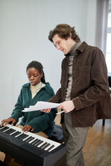 African young woman sitting at synthesizer and discussing the sheet music with her teacher during...