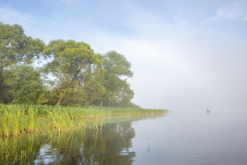 Obraz premium Mystical landscape. Fog in the early morning on the river. The trees near the water are illuminated by the rays of the rising sun.