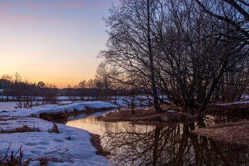 Naklejka premium Evening spring landscape, trees by the river, snow and thawed patches. Charming landscape in early spring against the backdrop of sunset. Melting ice and snow.