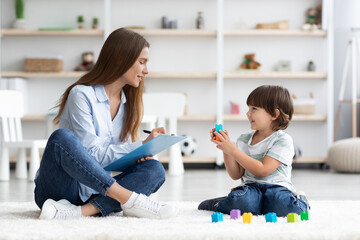 Cute little boy playing with colorful blocks, young professional woman kids psychologist writing...