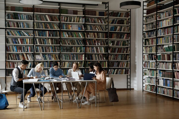 Group of multi racial students sit at table in spacious room of high school library, doing...