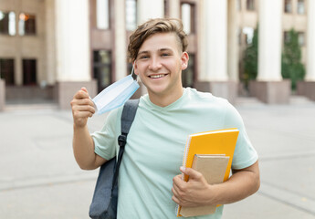 Happy student guy removing medical mask, posing outdoors with backpack and books, standing near...
