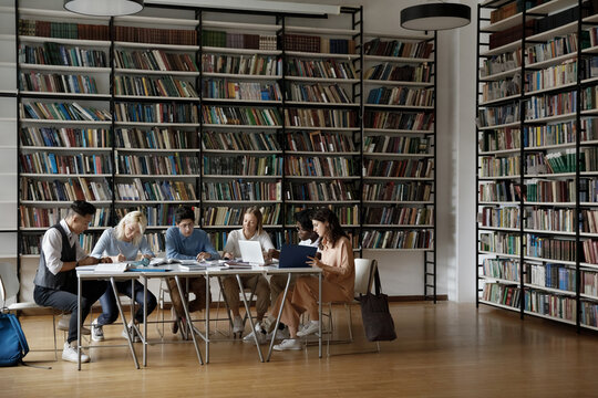 Group of multi racial students sit at table in spacious room of high school library, doing assignment, prepare for exams or admission looking focused. University education, studying process concept