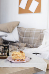 Fototapeta na wymiar Selective focus on a cake with a candle on a table set with a linen napkin next to a teapot with brewed tea and a cup, in the background a sofa with a checkered plaid against the wall with a mockup.