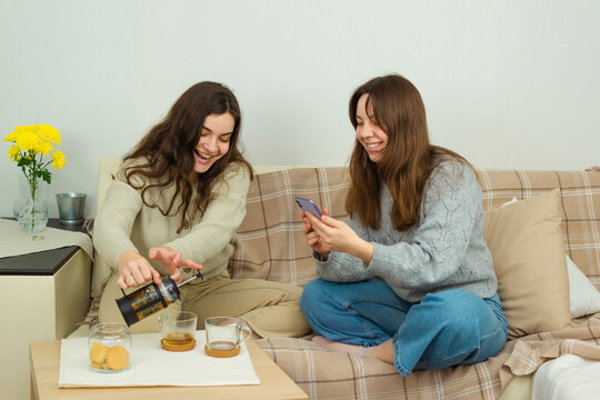 Two playful happy young women are sitting on the sofa and having fun, one smiles broadly and pours tea into a cup, the other laughs and takes pictures on a mobile phone camera.
