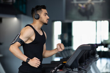 Fototapeta na wymiar Muscular Arab Male Athlete Wearing Wireless Headphones Running On Treadmill At Gym