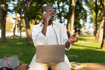 Excited african american male student talking on cellphone and using laptop, sitting in park or university campus