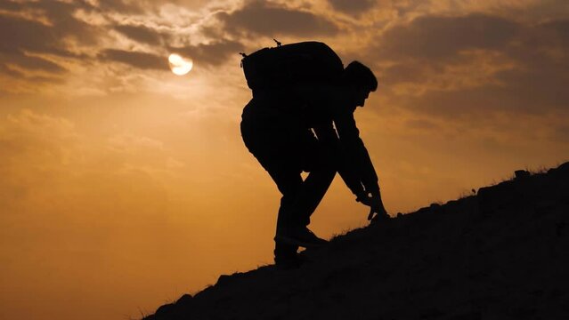 Silhouette shot of a hiker climbing the steep hill during the trek in front of the sun during sunset with clouds. Trekker struggling to climb the peak and slips. Adventure background.