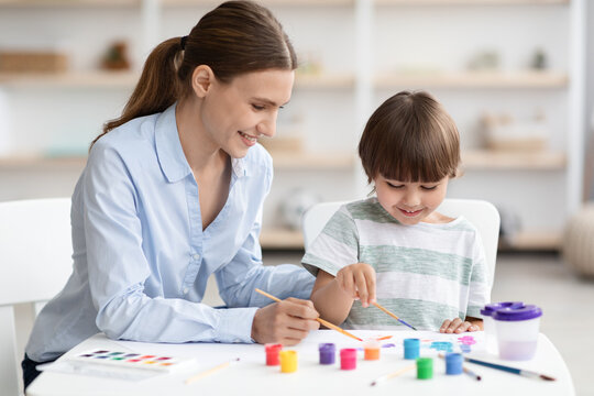 Cute little boy and his young mother drawing together with colorful paints, sitting at classroom and smiling