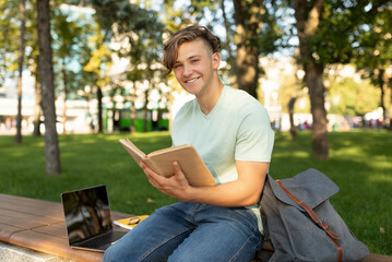 Fototapeta premium Happy student guy reading interesting book, having rest after classes, sitting on bench in parkland on sunny day