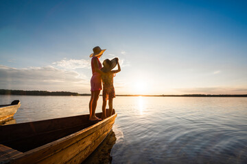 Mother and daughter stand together hugging on the bow of a wooden boat and enjoy the sunset by the lake or sea.