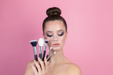 Makeup of a girl's face close-up in the studio on a pink background in the hands of a brush and paint. A young girl does makeup in the studio, paints eyelashes, eyebrows, lips look in the mirror.