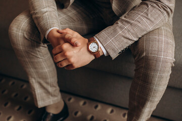 Man in a stylish suit with a wristwatch sits and holds his hands together. Groom getting ready in the morning before wedding ceremony. Man sit 