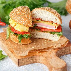 Gourmet homemade hamburger filled with slices of pork ham, cheese, tomato, lettuce, ketchup and eggs omelet on a wheat bun with sesame seeds on a wooden board. Delicious Breakfast. Selective focus