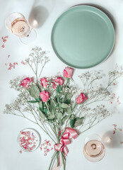 Flowers bunch with pink roses , empty plate and rose wine in glasses in sunlight on white background. Top view. Aesthetic flat lay