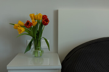 A bouquet of yellow and red tulips in a transparent vase on the bedside table in a bright bedroom