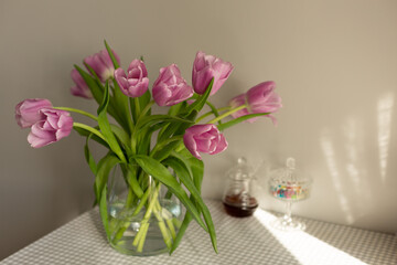 tulips of very peri color in a vase on the kitchen table, the table is covered with a gray checkered tablecloth, a glass candy bowl and a jar of dark honey are on the table