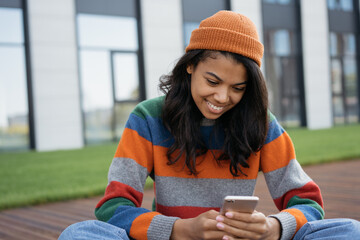 Smiling African American teenager wearing stylish hipster hat using mobile phone text messaging...
