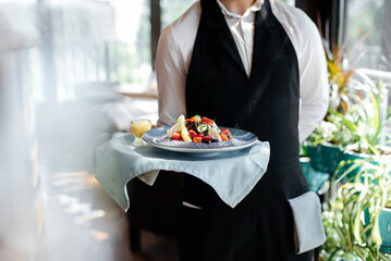 Close-up of a young waiter in a stylish uniform carrying an exquisite salad to a client in a...