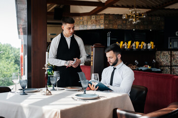 A young businessman in a fine restaurant examines the menu and makes an order to a young waiter in a stylish apron. Customer service. Table service in the restaurant.