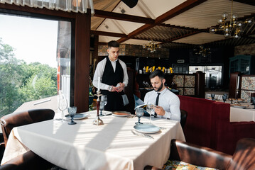 A young businessman in a fine restaurant examines the menu and makes an order to a young waiter in a stylish apron. Customer service. Table service in the restaurant.