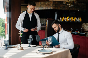 A young businessman in a fine restaurant examines the menu and makes an order to a young waiter in a stylish apron. Customer service. Table service in the restaurant.