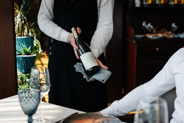 A young sommelier in a stylish uniform demonstrates and offers the client fine wine in the restaurant. Customer service. Table setting in a fine restaurant. Close-up. - obrazy, fototapety, plakaty