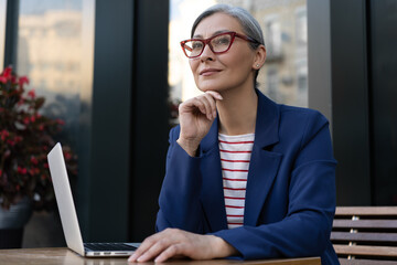 Pensive mature businesswoman using laptop computer working online at workplace. Portrait of middle aged grey haired manager wearing stylish eyeglasses planning startup. Successful business