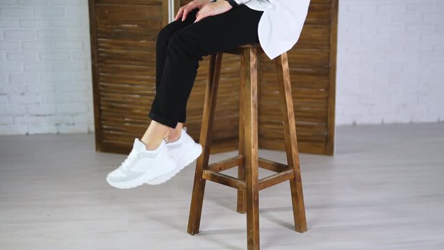 Woman sits on a high stool waving her feet in white sneakers. Model jumps off the tabouret and walks away in comfortable footwear.
