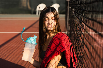 Athletic fit girl drinks water from a bottle. Rest after training on the tennis court.