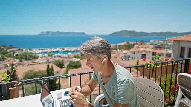 dolly zoom footage man in erphones working witth laptop outdoors by beautiful blue sea view, speaking by online video call. Concept remote work and travel.